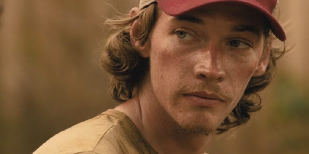 Close-up of Cooper Norris in Landman, wearing a red cap and looking intense while standing outdoors in the Texas oilfields.