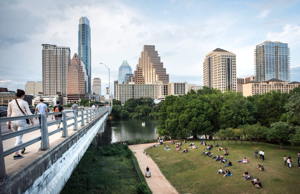 Modern downtown Austin skyline with pedestrians crossing bridge, contrasting Landman's gritty oilfields with sleek corporate cityscapes.